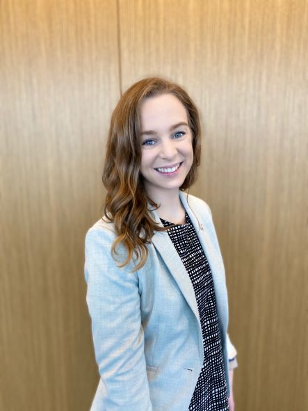 woman in a gray blazer standing in front of a brown wooden wall.